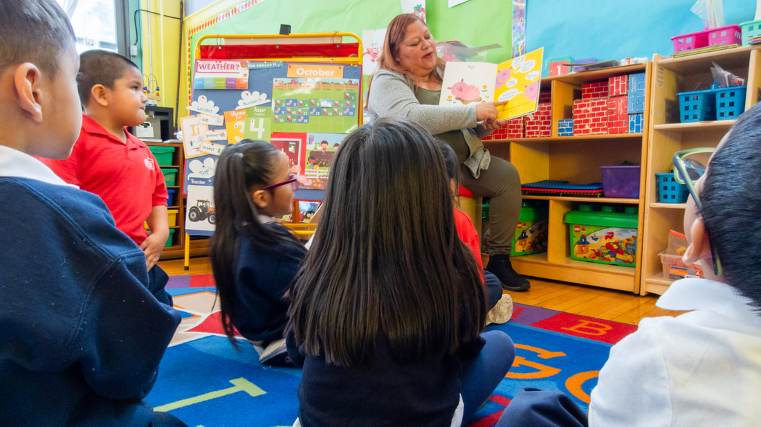 Mrs. Gutierrez, the Pre-K 3 & 4 teacher sits in front of the class reading out loud from a book. The students sit on a brightly colored carpet in front of her.