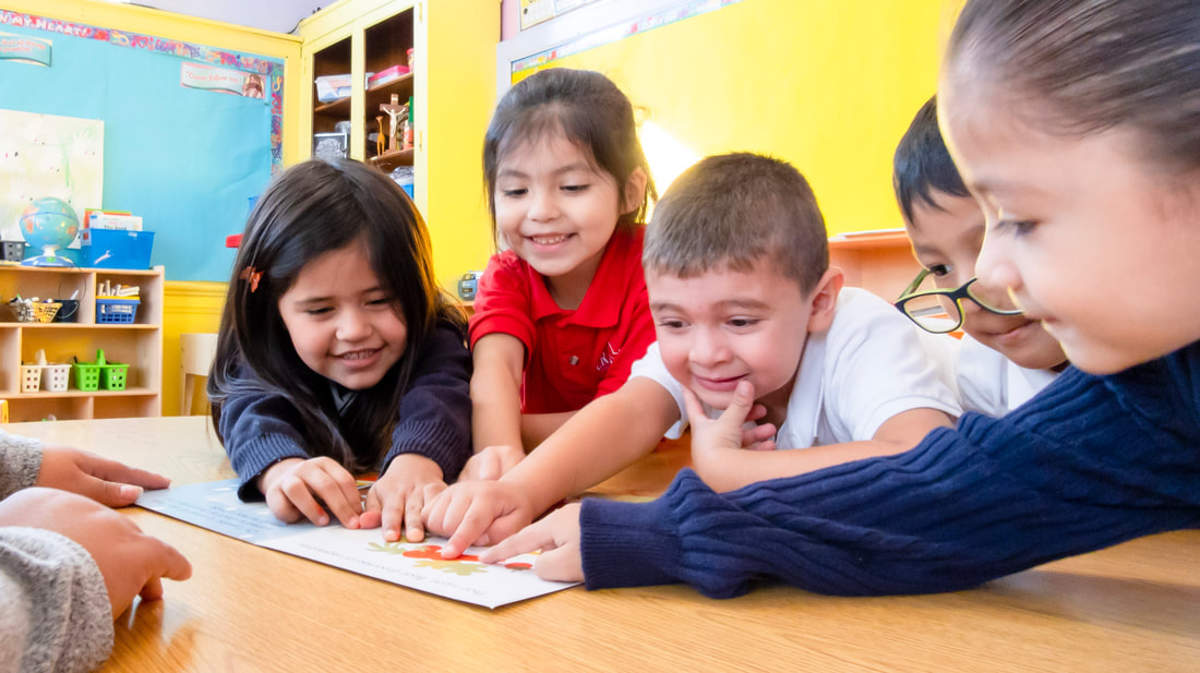 Three female and two male early childhood students smile and point at a picture while sitting around a table.