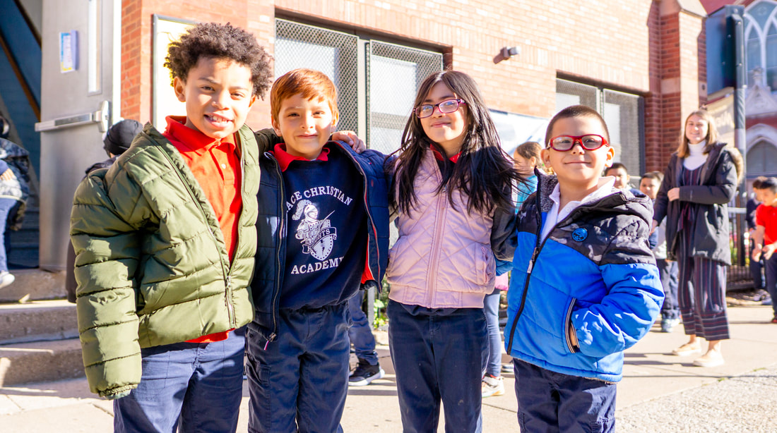 Three male and one female elementary school students smiling in front of the school on a sunny day