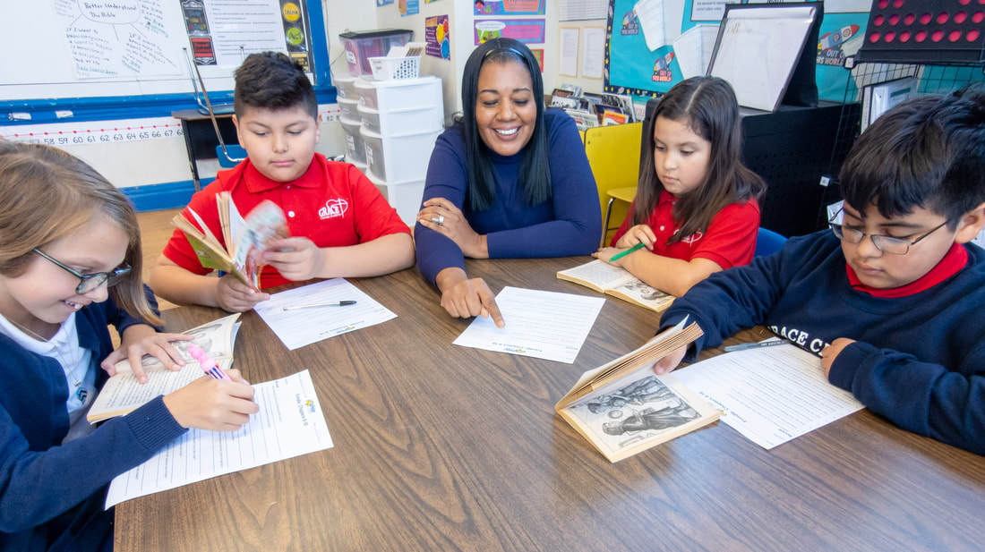 Mrs. Segal sitting at a table with two female and two male elementary school students in uniform. Mrs. Segal is smiling and instructing while pointing at a piece of paper on the table. The students are each holding a book.