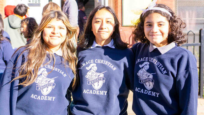 Three female middle schools students smiling with their arms around each other on a sunny day in front of the school. They are all wearing blue Grace Christian Academy sweatshirts.