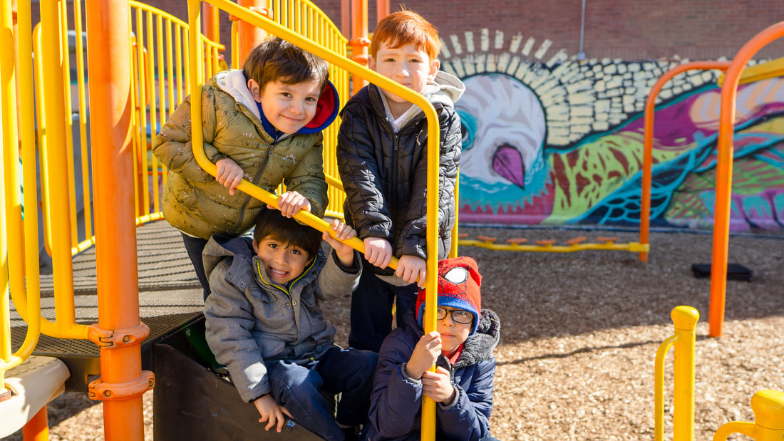 Four male students smile on the steps of playground equipment on a sunny day. A brightly colored mural is displayed in the background on the school building.