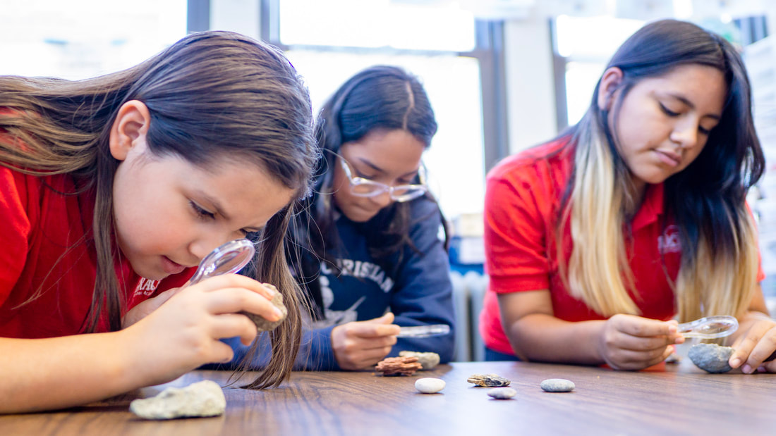 Three female middle school students in uniform sit at a table inspecting rocks with a magnifying glass.